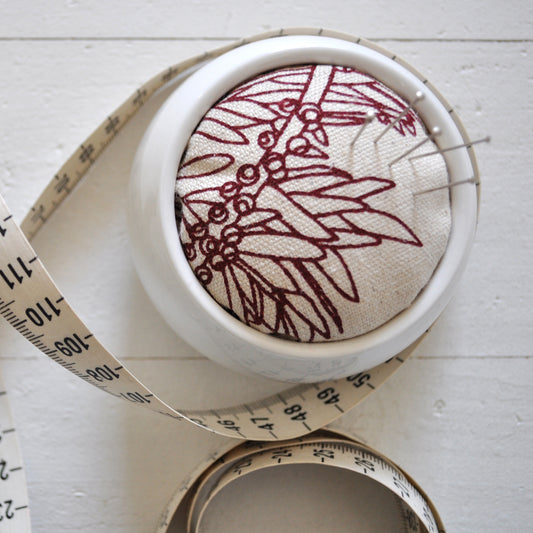Pinch River ceramic pin cushion featuring Bottle Brush print in Wine Berry (colourway 1). Close-up top view. Product image is styled with a measuring tape and pins.