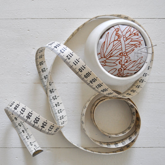Pinch River ceramic pin cushion featuring Bottle Brush print in Burnt Orange (colourway 2). Top view. Product image is styled with a measuring tape and pins.
