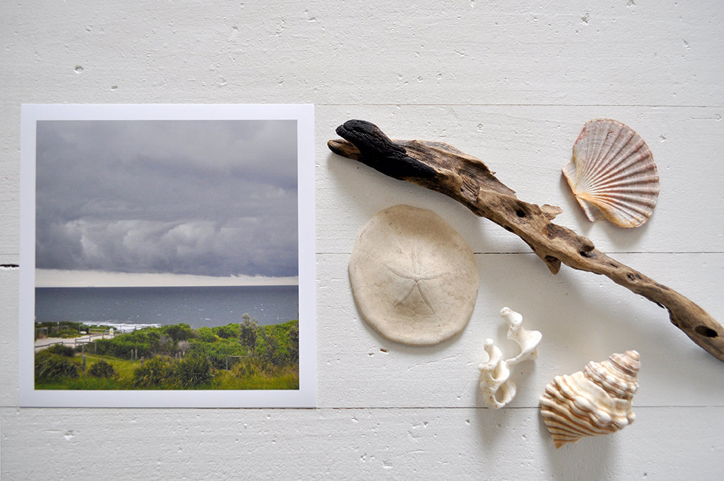 Pinch River unframed 17cm x 17cm Photographic Print on Paper Titled: Storm Clouds. The subject of the print is storm clouds over the ocean. Top view with sea shells and driftwood next to it to style the product image.
