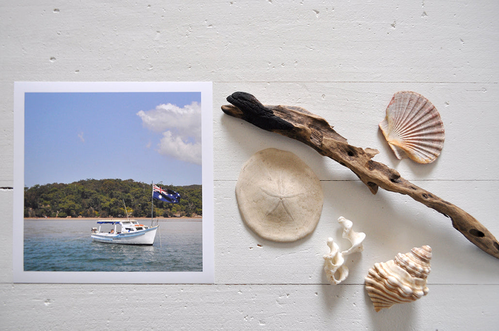Pinch River unframed 17cm x 17cm Photographic Print on Paper Titled: Spirit of Australia. The subject of the print is a boat on the water flying the Australian flag. Top view with sea shells and driftwood next to it to style the product image.