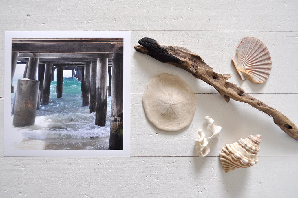 Pinch River unframed 17cm x 17cm Photographic Print on Paper Titled: Underneath the Jetty #2. The subject of the print is a close-up of the iconic Catherine Hill Bay Jetty from underneath, with the weather-worn timber pilings, waves rolling in and beautiful aqua blue of the water in the background. Top view with sea shells and driftwood next to it to style the product image.