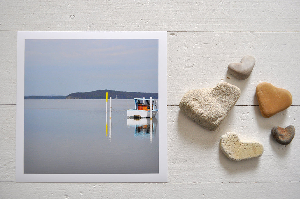Photographic Print on Paper Titled: Nautical Reflections. The subject of the print is a boat docked at a marina and reflections on the still lake water. Top view with love heart shaped stones to style the product image.