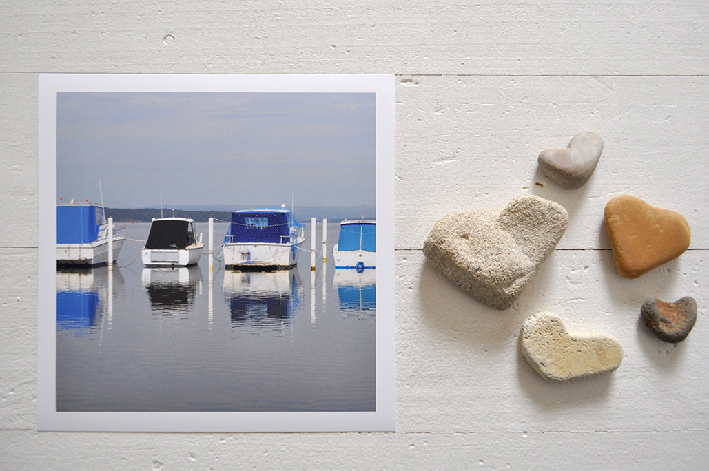 Pinch River unframed 17cm x 17cm Photographic Print on Paper Titled: Lakeside Harmony. The subject of the print is 4 boats docked at a marina all with blue canopies and reflections on the still lake water. Top view with love heart shaped stones to style the product image.
