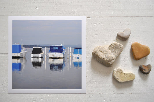 Pinch River unframed 17cm x 17cm Photographic Print on Paper Titled: Lakeside Harmony. The subject of the print is 4 boats docked at a marina all with blue canopies and reflections on the still lake water. Top view with love heart shaped stones to style the product image.