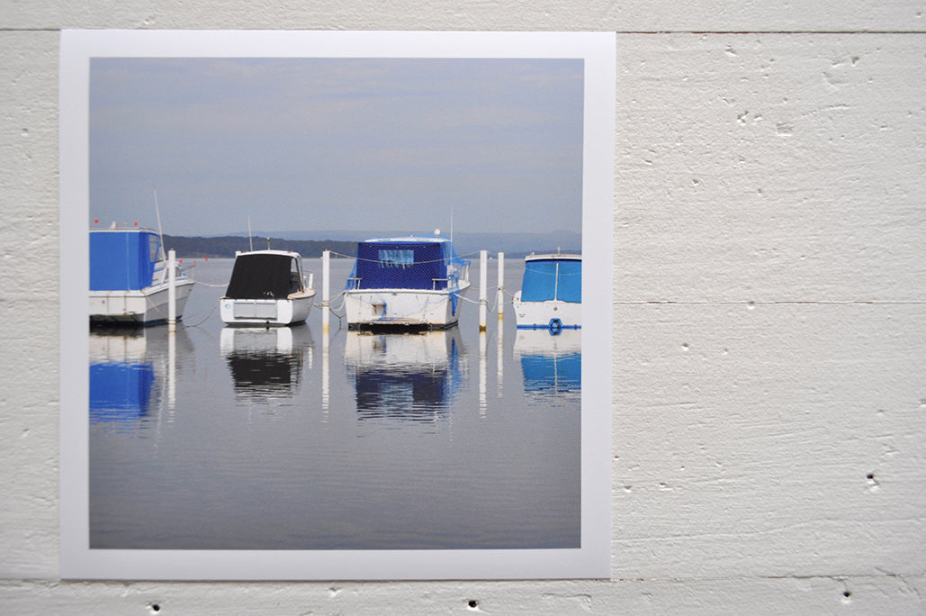 Photographic Print on Paper Titled: Lakeside Harmony. The subject of the print is 4 boats docked at a marina all with blue canopies and reflections on the still lake water. Top view.