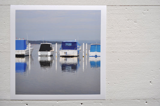 Photographic Print on Paper Titled: Lakeside Harmony. The subject of the print is 4 boats docked at a marina all with blue canopies and reflections on the still lake water. Top view.