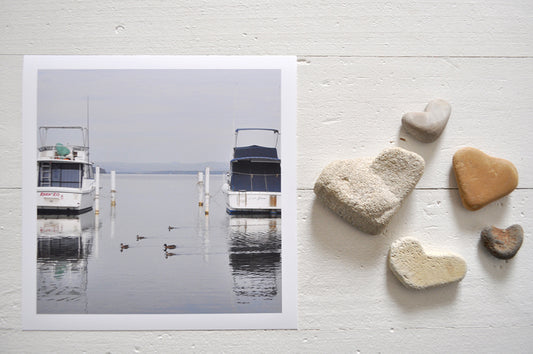 Pinch River unframed 17cm x 17cm Photographic Print on Paper Titled: Lakeside Cruising #1. The subject of the print is ducks and boats docked at a marina with reflections on the still lake water. Top view with love heart shaped stones to style the product image.