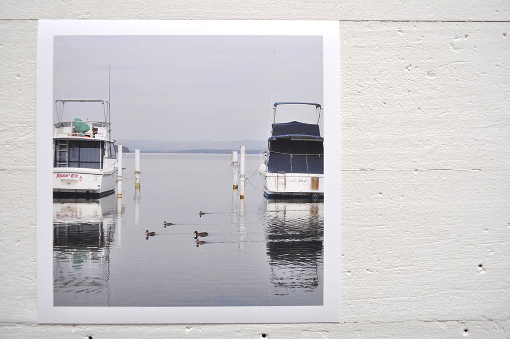 Pinch River unframed 17cm x 17cm Photographic Print on Paper Titled: Lakeside Cruising #1. The subject of the print is ducks and boats docked at a marina with reflections on the still lake water. Top view.
