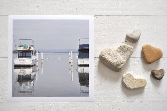 Pinch River unframed 17cm x 17cm Photographic Print on Paper Titled: Lakeside Cruising #2. The subject of the print is pelicans and boats docked at a marina with reflections on the still lake water. Top view with love heart shaped stones to style the product image.
