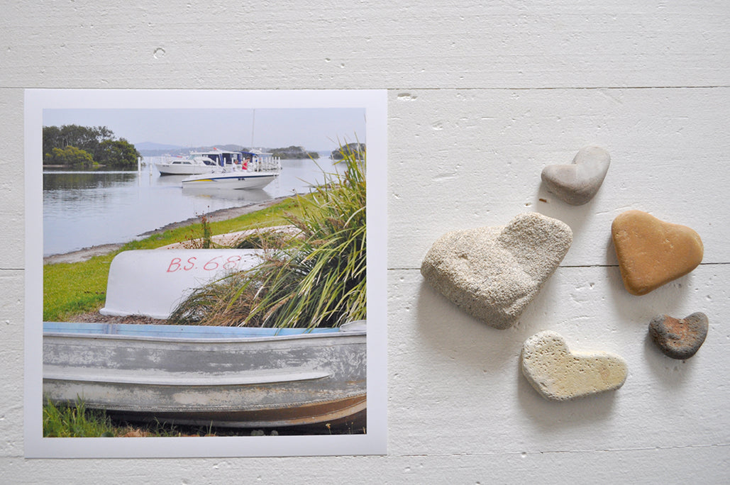 Pinch River unframed 17cm x 17cm Photographic Print on Paper Titled: Tinnies on the Shore. The subject of the print is a couple of tinnies on the lake shore with boats in the water in the background. Top view with love heart shaped stones to style the product image.