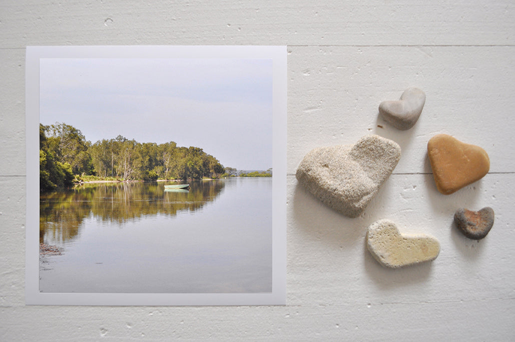 Pinch River unframed 17cm x 17cm Photographic Print on Paper Titled: The Green Boat. The subject of the print is a green row boar on the lake, with reflections of the treed shoreline on the still lake water. Top view with love heart shaped stones to style the product image.