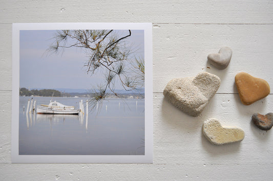 Pinch River unframed 17cm x 17cm Photographic Print on Paper Titled: Boats on the Lake #1. The subject of the print a couple of boats moored on the lake with a She Oak branch in the foreground. Top view with love heart shaped stones to style the product image.