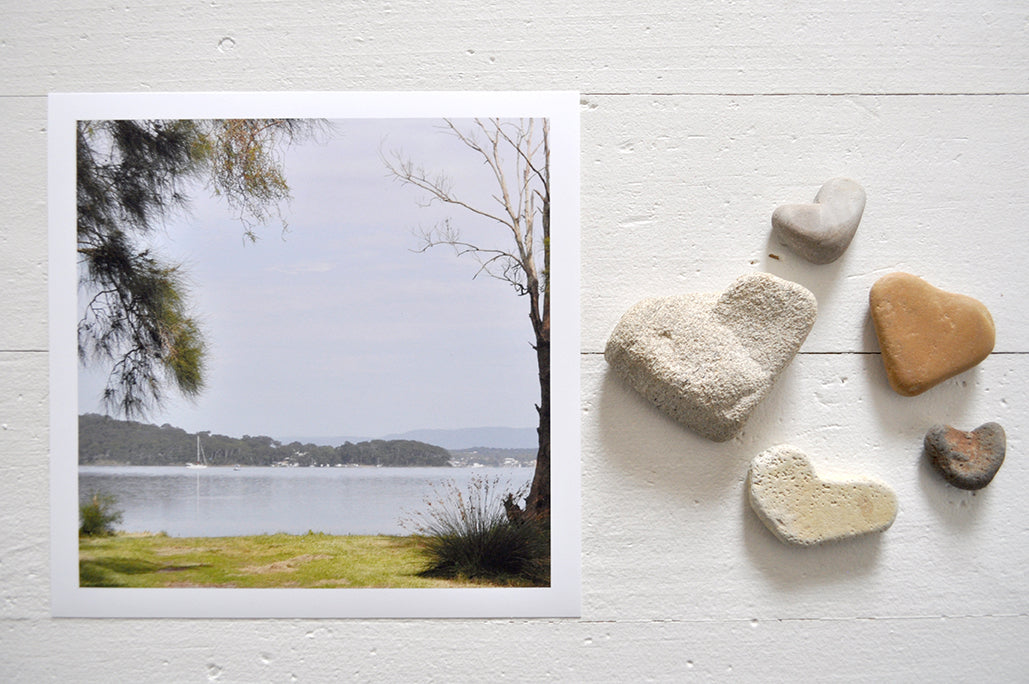 Pinch River unframed 17cm x 17cm Photographic Print on Paper Titled: Lakeside Vista. The subject of the print is a vista of the lake framed by trees on either side. Top view with love heart shaped stones to style the product image.
