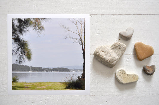 Pinch River unframed 17cm x 17cm Photographic Print on Paper Titled: Lakeside Vista. The subject of the print is a vista of the lake framed by trees on either side. Top view with love heart shaped stones to style the product image.