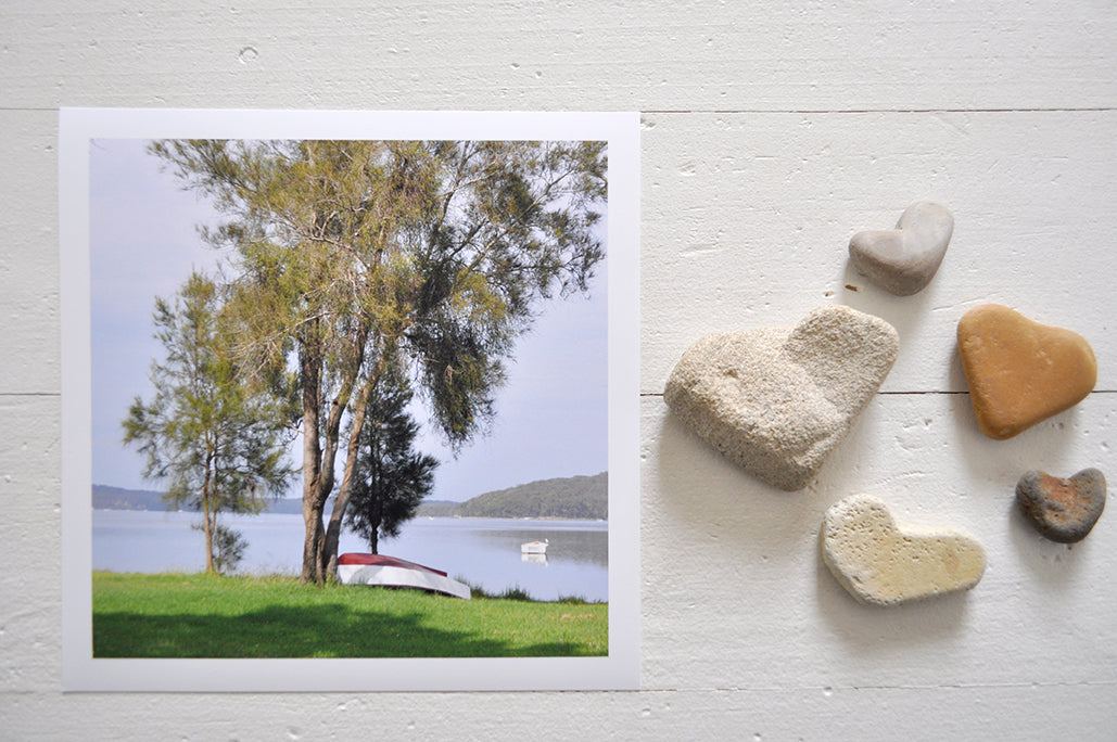 Pinch River unframed 17cm x 17cm Photographic Print on Paper Titled: Waiting for Sunday #1. The subject of the print is a large tree on the lake shore with an upturned row boat underneath it. Top view with love heart shaped stones to style the product image.