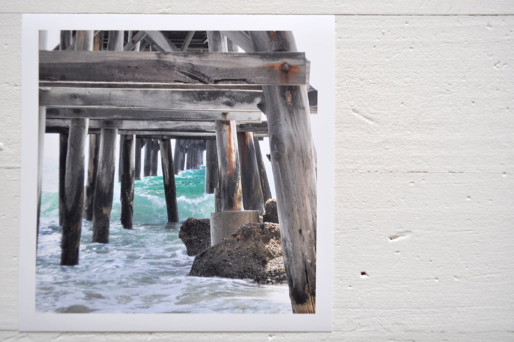 Pinch River unframed 17cm x 17cm Photographic Print on Paper Titled: Underneath the Jetty #1. The subject of the print is a close-up of the iconic Catherine Hill Bay Jetty from underneath, with the weather-worn timber pilings, waves rolling in and beautiful aqua blue of the water in the background. Top view.