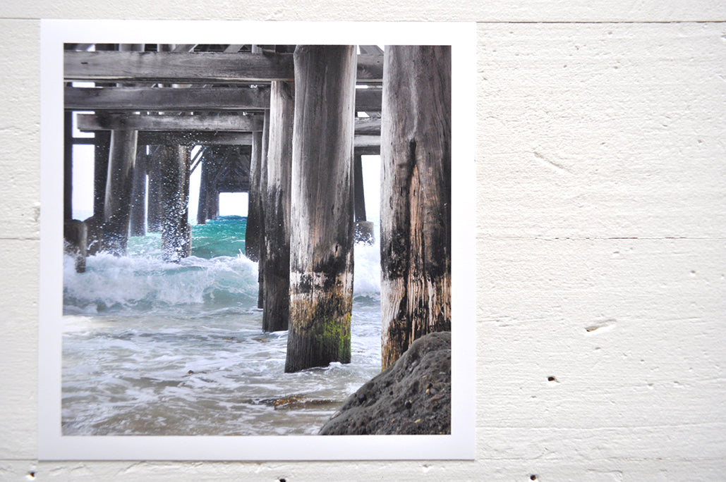 Pinch River unframed 17cm x 17cm Photographic Print on Paper Titled: Underneath the Jetty #3. The subject of the print is a close-up of the iconic Catherine Hill Bay Jetty from underneath, with the weather-worn timber pilings, waves crashing and beautiful aqua blue of the water in the background. Top view.