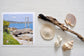 Pinch River unframed 17cm x 17cm Photographic Print on Paper Titled: Reids Reserve #1. The subject of the print is a coastal rocky outcrop looking out over the ocean, with mooring posts in the background looking towards the lake's entrance. Top view with sea shells and driftwood next to it to style the product image.