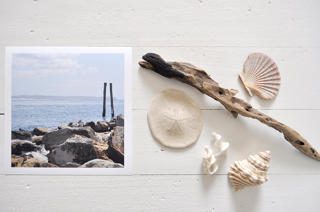 Pinch River unframed 17cm x 17cm Photographic Print on Paper Titled: Reids Reserve #2. The subject of the print is a coastal rocky outcrop looking out over the ocean, with mooring posts in the background. Top view with sea shells and driftwood next to it to style the product image.