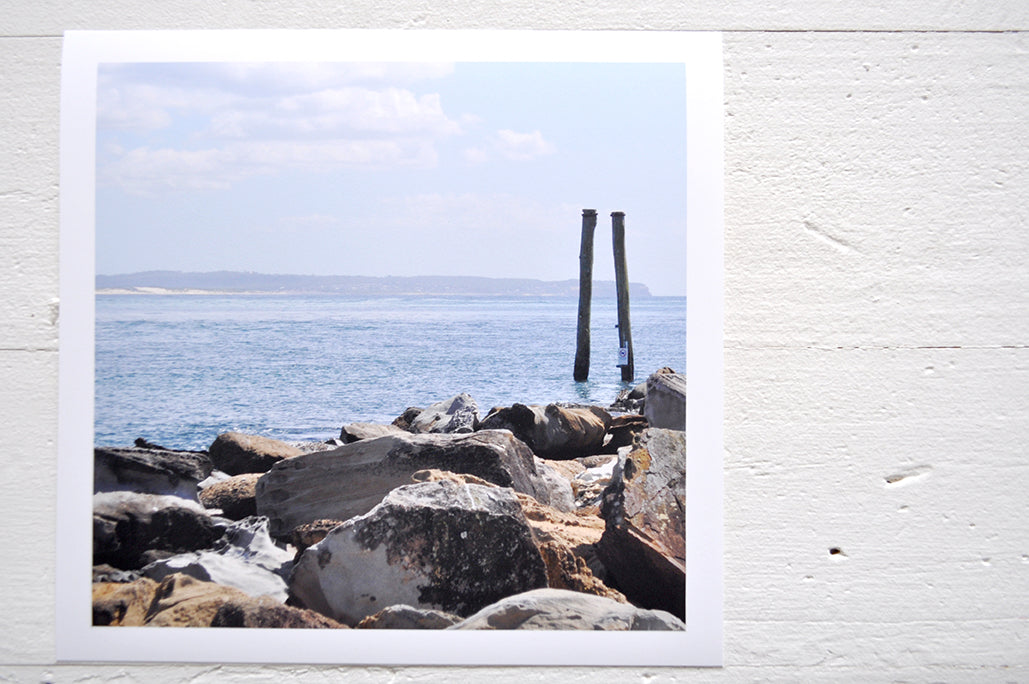 Pinch River unframed 17cm x 17cm Photographic Print on Paper Titled: Reids Reserve #2. The subject of the print is a coastal rocky outcrop looking out over the ocean, with mooring posts in the background. Top view.