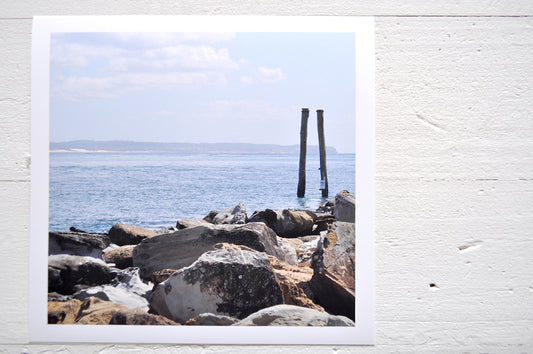 Pinch River unframed 17cm x 17cm Photographic Print on Paper Titled: Reids Reserve #2. The subject of the print is a coastal rocky outcrop looking out over the ocean, with mooring posts in the background. Top view.