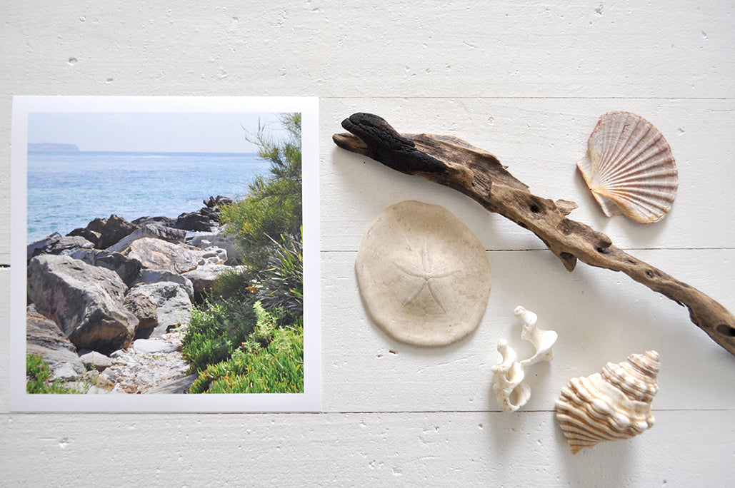 Pinch River unframed 17cm x 17cm Photographic Print on Paper Titled: Reids Reserve #3. The subject of the print is a coastal rocky outcrop looking out over the ocean. Top view with sea shells and driftwood next to it to style the product image.