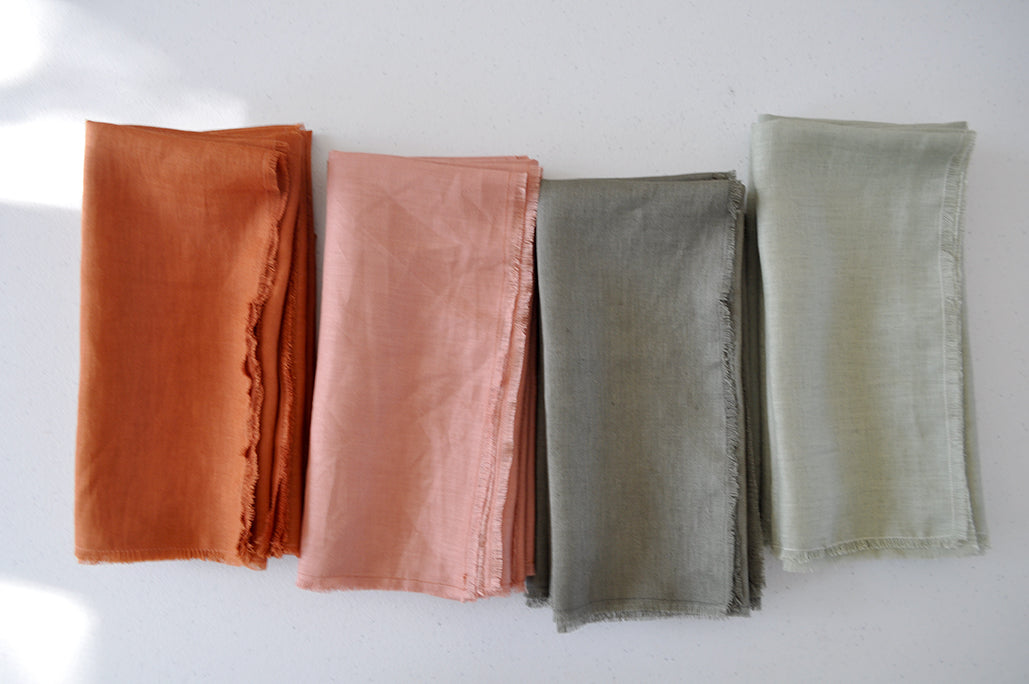 Assorted Pinch River pure linen napkins, folded, grouped by colour: Rust, Coral, Olive, Sage. Top view.