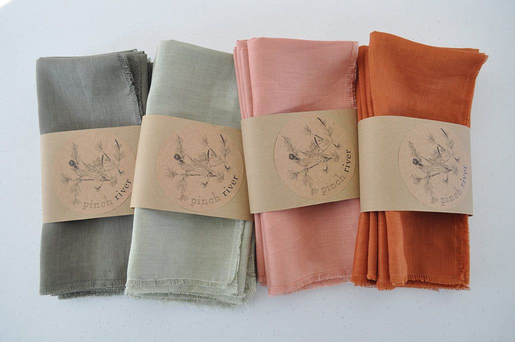 Assorted Pinch River pure linen napkins, folded, grouped by colour: Rust, Coral, Olive, Sage with branded belly bands / products sleeves. Top view.
