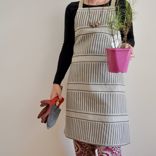 Model wearing Pinch River bib apron / full apron in Corduroy Pathway print holding a potted plant and gardening tools against a plain background. Front view. Pose #1.