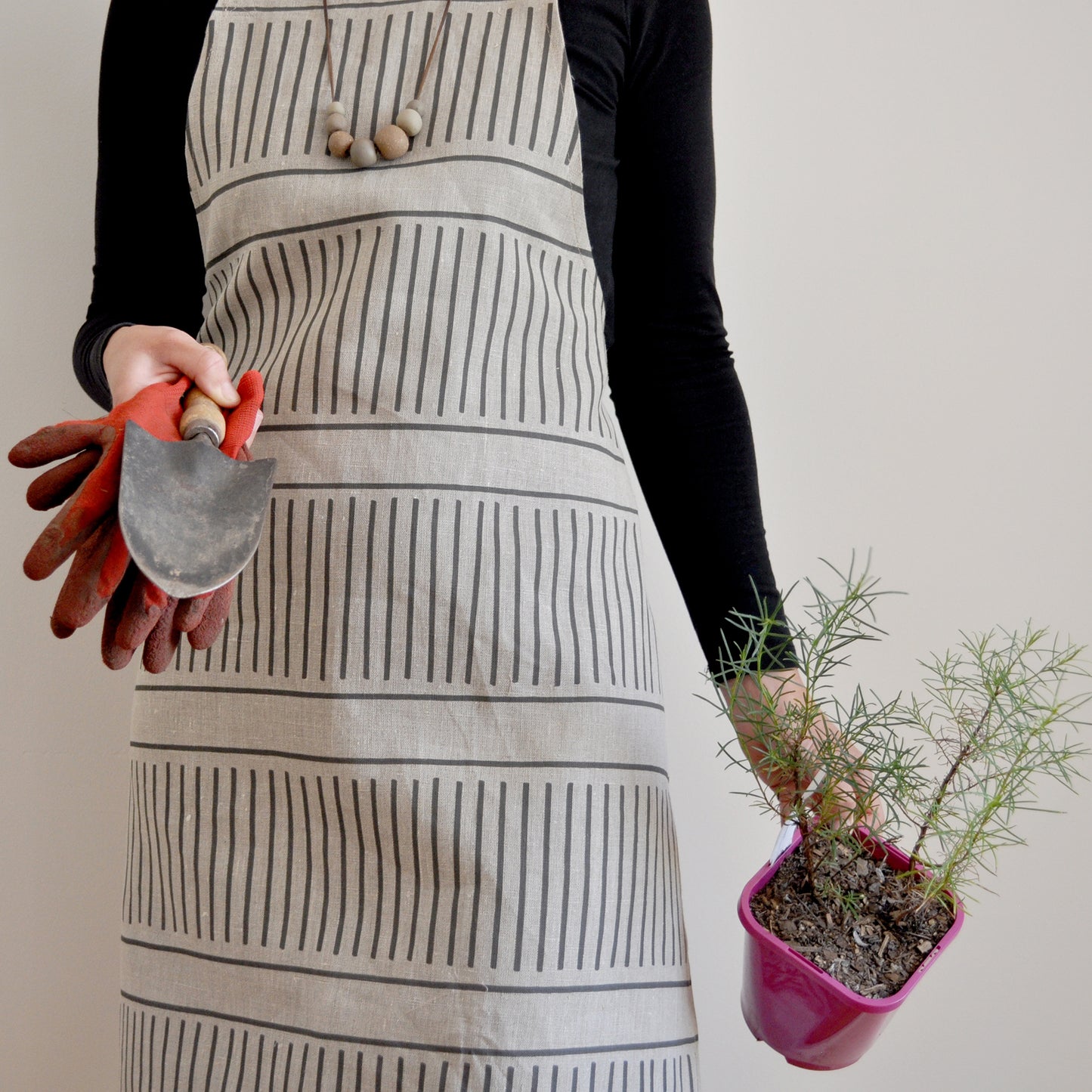 Model wearing Pinch River bib apron / full apron in Corduroy Pathway print holding a potted plant and gardening tools against a plain background. Front view. Pose #2.