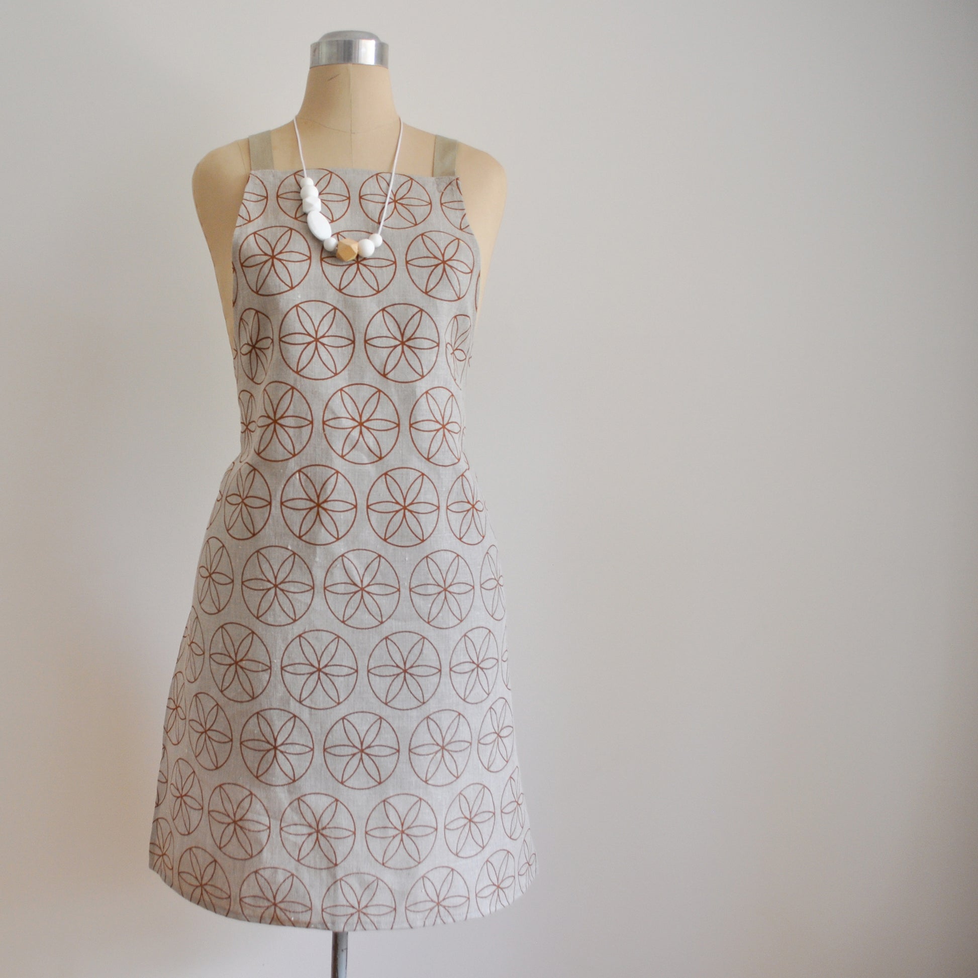 Pinch River bib apron / full apron in Flower of Life print, Burnt Orange on Natural Linen (colourway 2) on dressmakers dummy against a plain background. Front View.