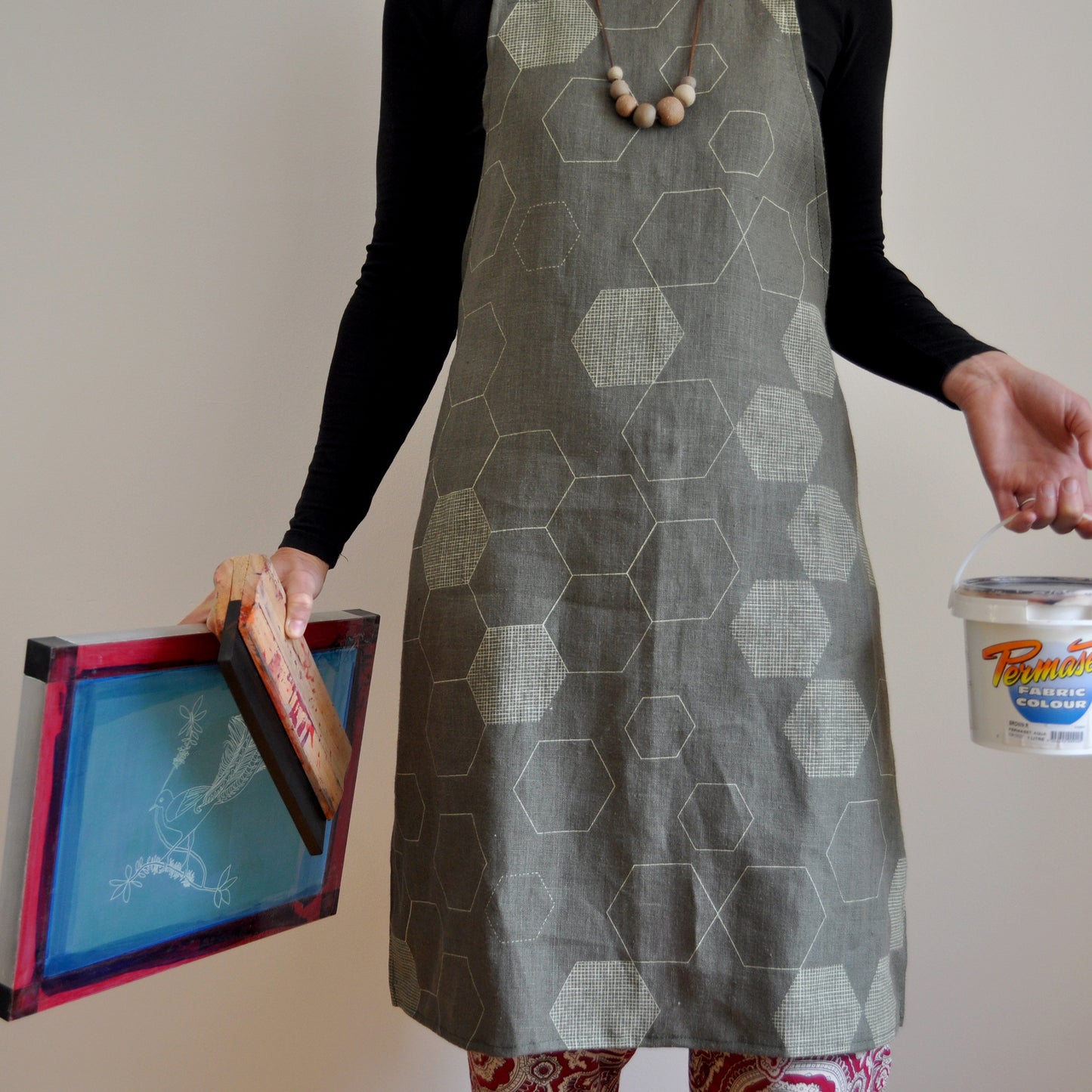 Model wearing Pinch River bib apron / full apron in Honeycomb print holding a a silk screen, squeegee and a tub of printing ink against a plain background. Front View.