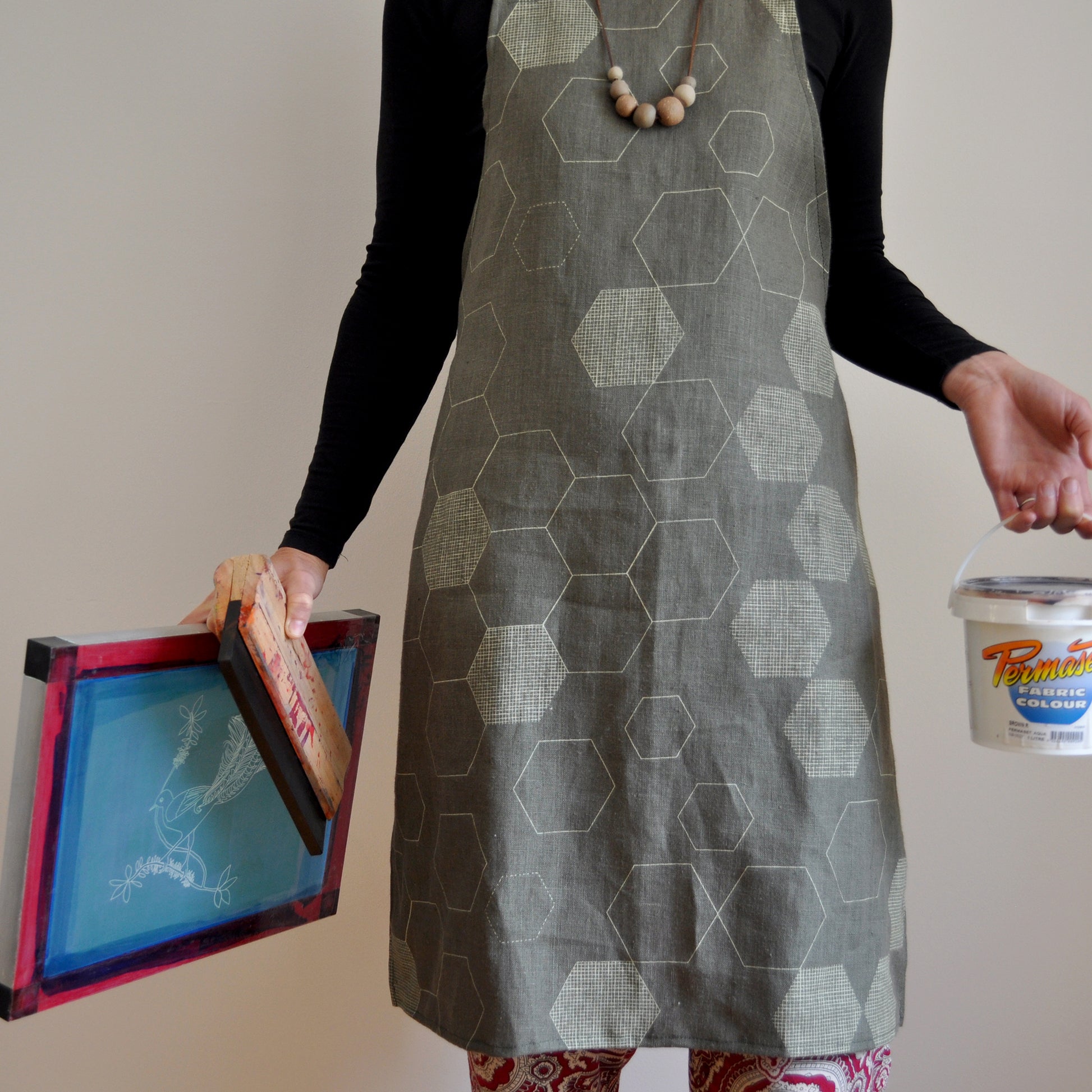 Model wearing Pinch River bib apron / full apron in Honeycomb print holding a a silk screen, squeegee and a tub of printing ink against a plain background. Front View.