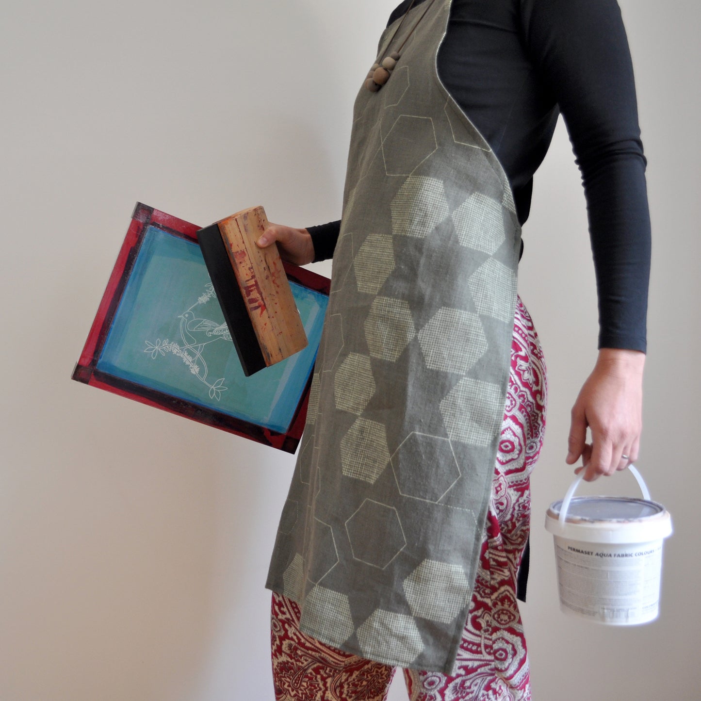 Model wearing Pinch River bib apron / full apron in Honeycomb print holding a a silk screen, squeegee and a tub of printing ink against a plain background. Side View.