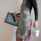 Model wearing Pinch River bib apron / full apron in Honeycomb print holding a a silk screen, squeegee and a tub of printing ink against a plain background. Side View.