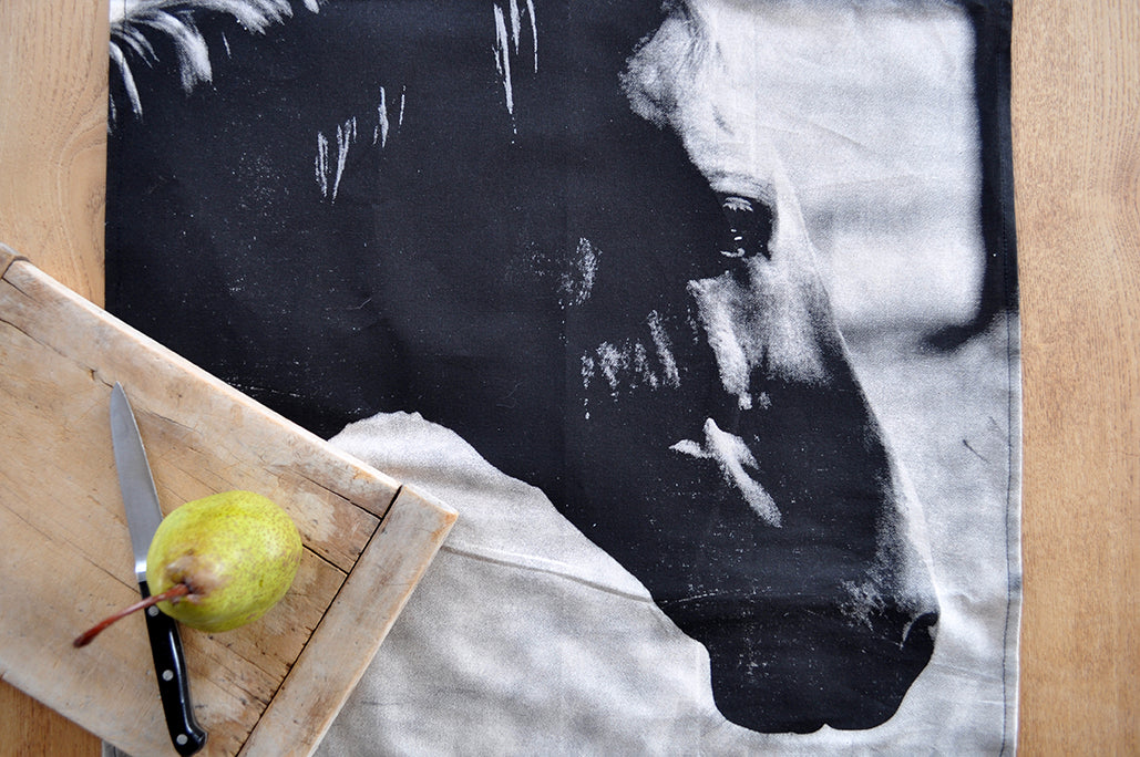 Pinch River handprinted halftone Horse tea towel. Top view. Image styled with vintage time-worn cutting board, pear and knife next to tea towel.