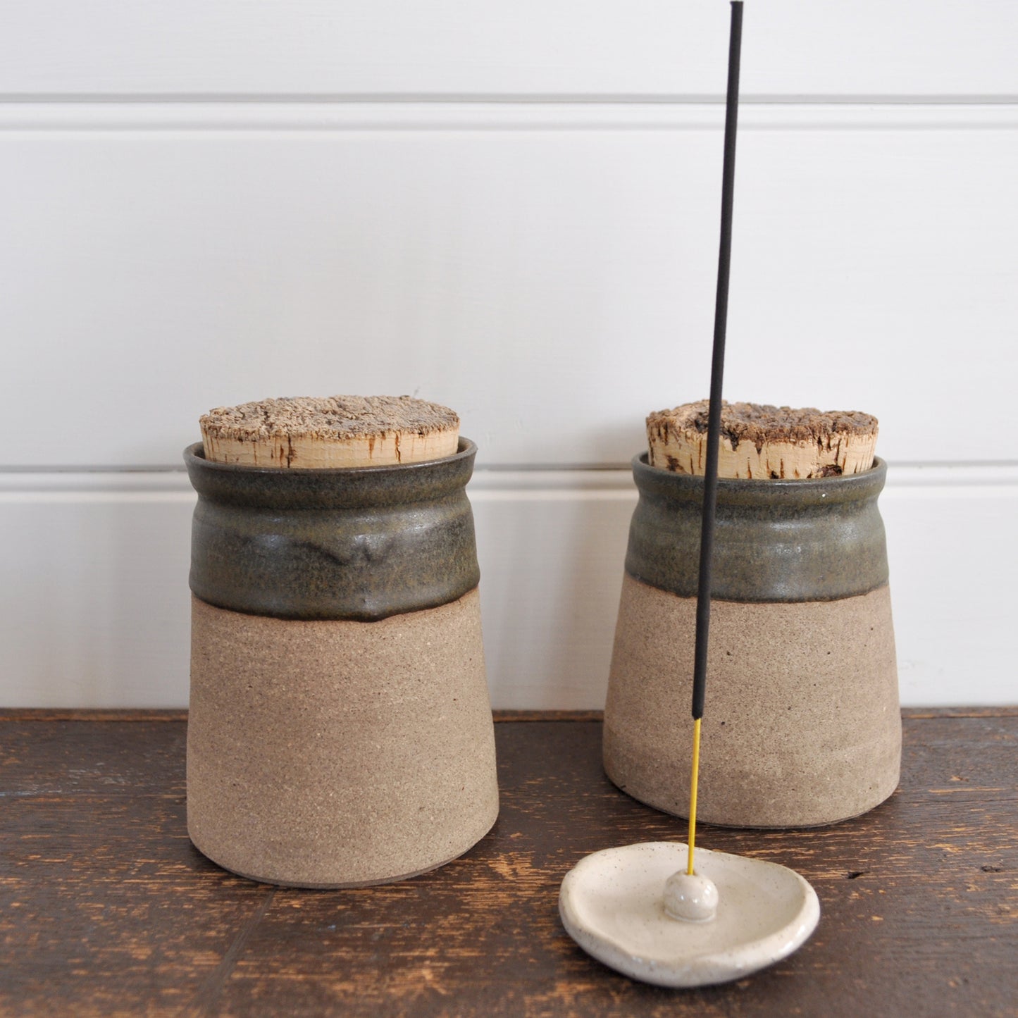 Handmade ceramic incense holder in foreground, with an incense stick in it. Product image is styled with two lidded ceramic pots in the back ground.