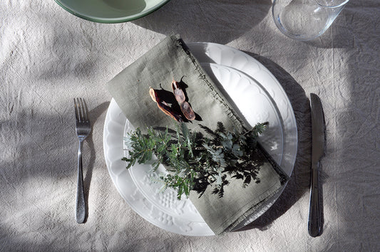 Dining table setting with a white plate, Pinch River pure linen napkin in Olive, and greenery on a linen tablecloth. Top view.