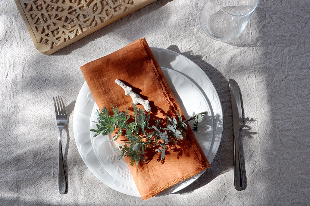Dining table setting with a white plate, Pinch River pure linen napkin in Rust, and greenery on a linen tablecloth. Top view. Landscape orientation.