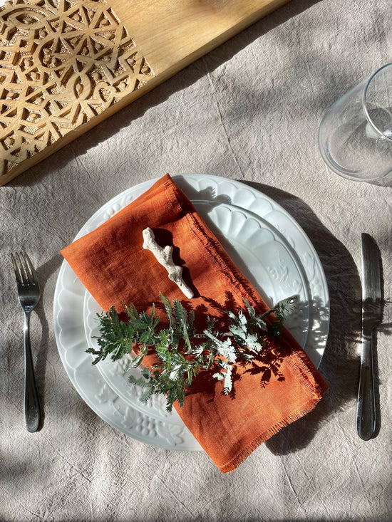 Dining table setting with a white plate, Pinch River pure linen napkin in Rust, and greenery on a linen tablecloth. Top view. Portrait orientation.