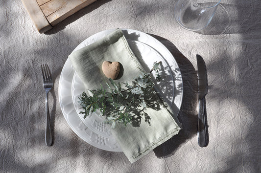 Dining table setting with a plate, Pinch River pure linen napkin in Sage, and cutlery on linen tablecloth.