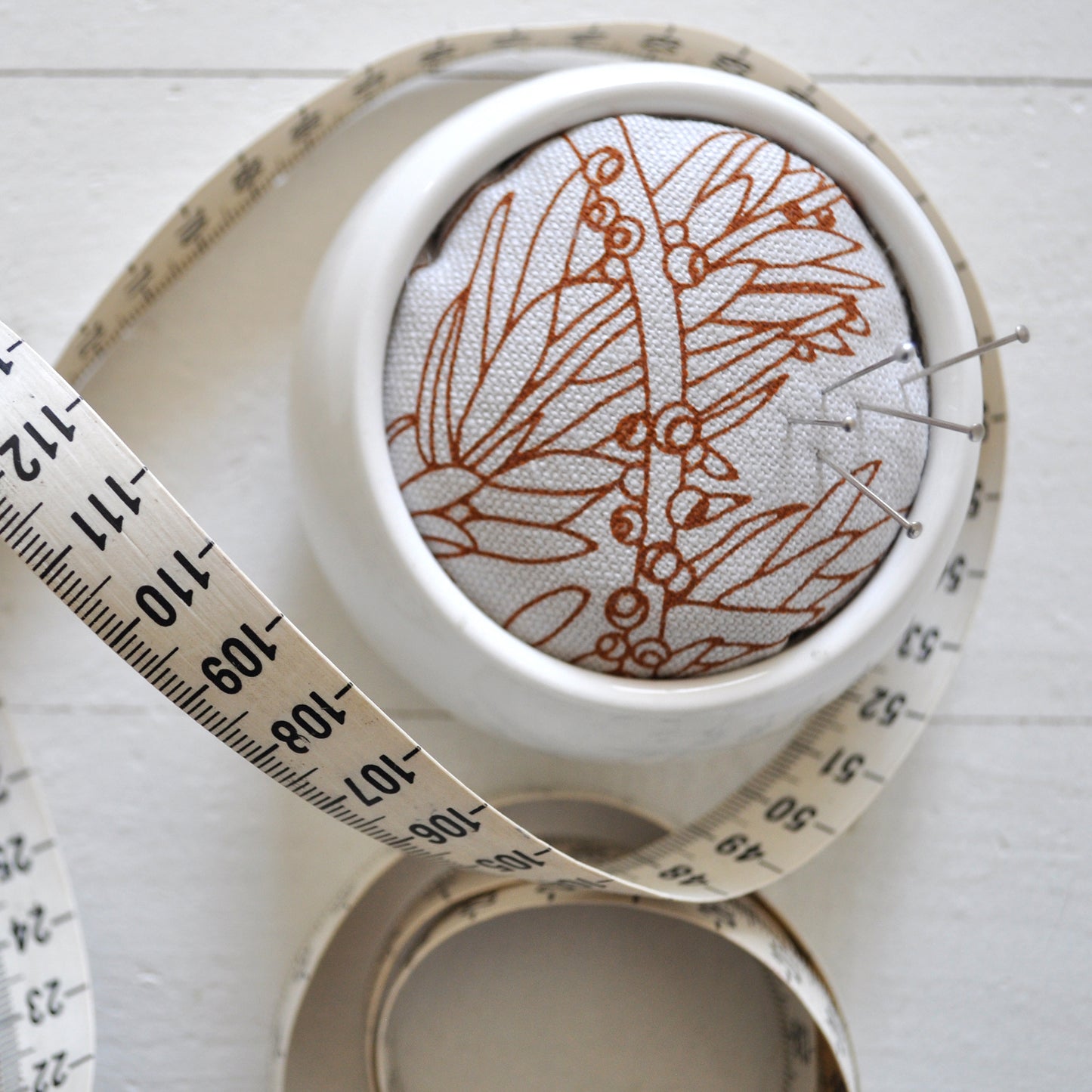Pinch River ceramic pin cushion featuring Bottle Brush print in Burnt Orange (colourway 2). Close-up top view. Product image is styled with a measuring tape and pins.
