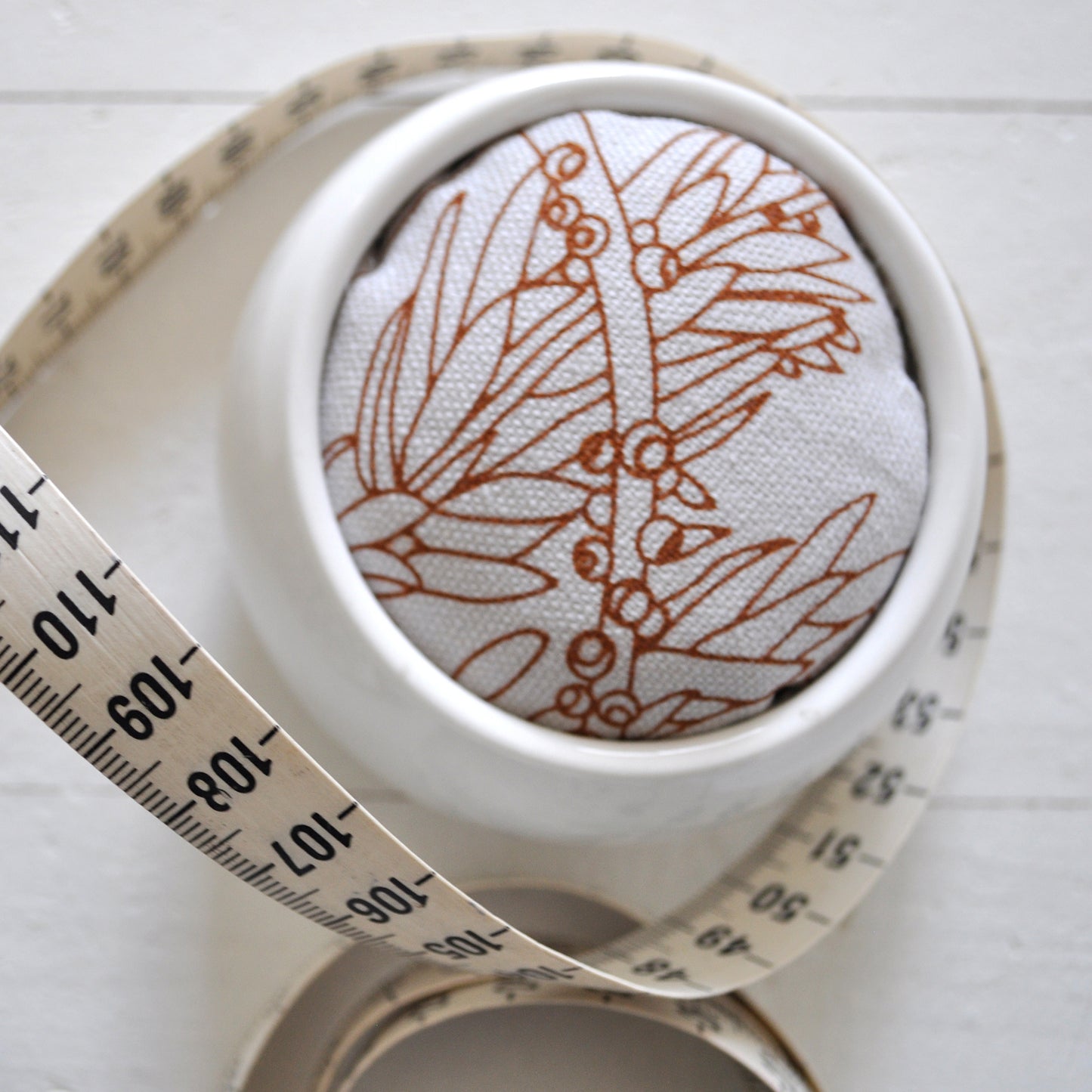 Pinch River ceramic pin cushion featuring Bottle Brush print in Burnt Orange (colourway 2). Close-up top view. Product image is styled with a measuring tape.