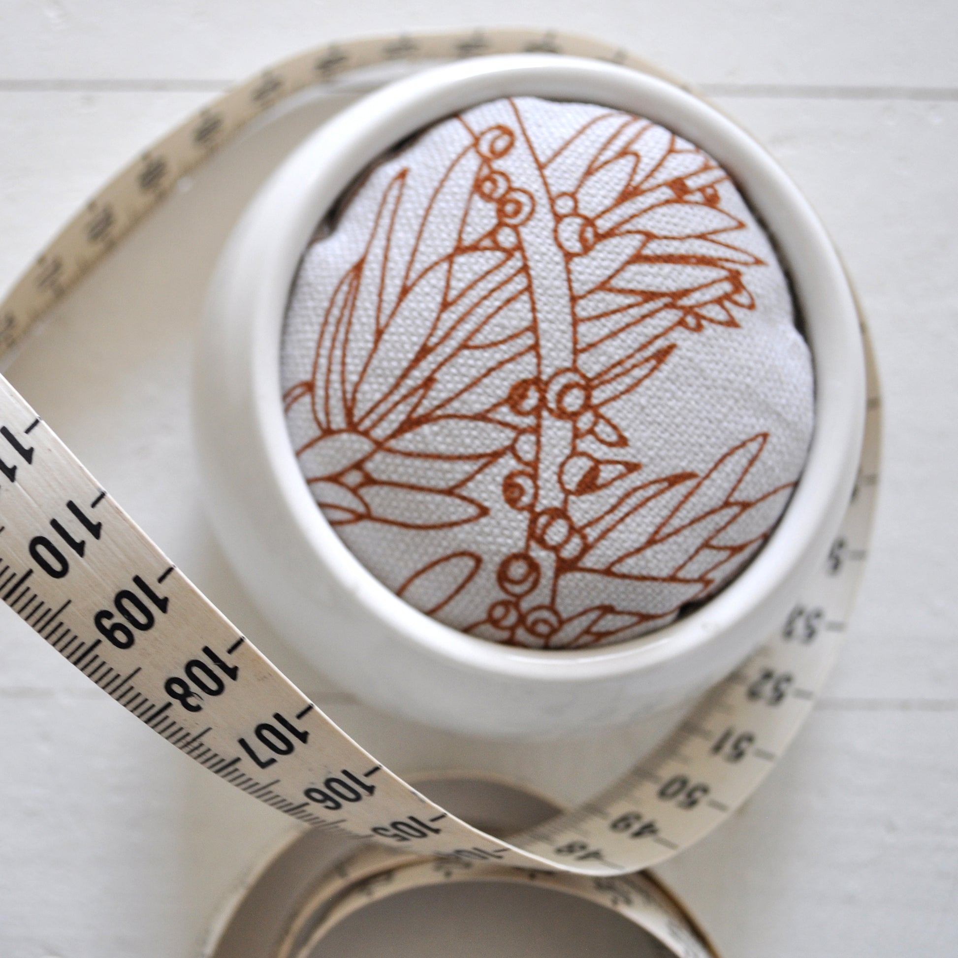 Pinch River ceramic pin cushion featuring Bottle Brush print in Burnt Orange (colourway 2). Close-up top view. Product image is styled with a measuring tape.