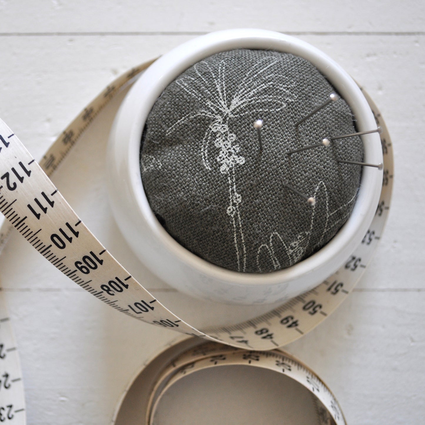 Pinch River ceramic pin cushion featuring Bottle Brush print in Moss Green (colourway 3). Close-up top view. Product image is styled with a measuring tape and pins.