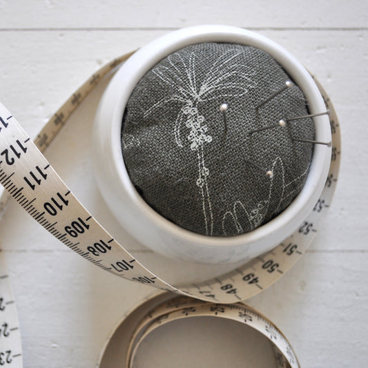 Pinch River ceramic pin cushion featuring Bottle Brush print in Moss Green (colourway 3). Close-up top view. Product image is styled with a measuring tape and pins.