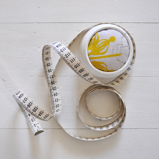 Pinch River ceramic pin cushion featuring Firewheel print in Buddha Gold (colourway 1_v1). Top view. Product image is styled with a measuring tape and pins.