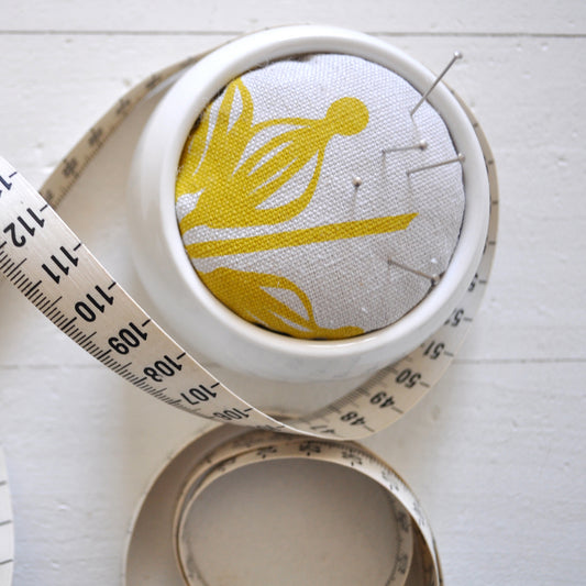 Pinch River ceramic pin cushion featuring Firewheel print in Buddha Gold (colourway 1_v1). Close-up top view. Product image is styled with a measuring tape and pins.