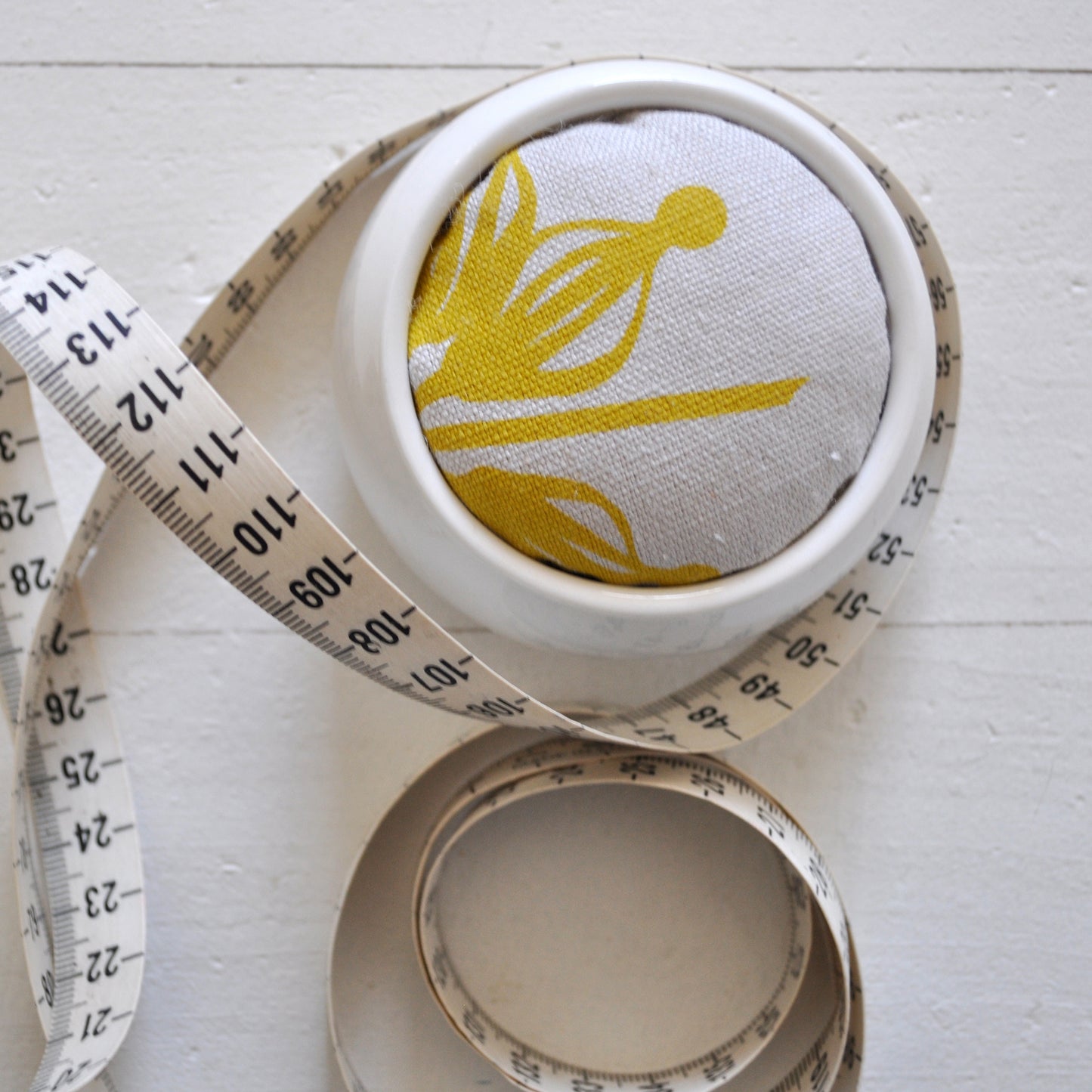 Pinch River ceramic pin cushion featuring Firewheel print in Buddha Gold (colourway 1_v1). Close-up top view. Product image is styled with a measuring tape.