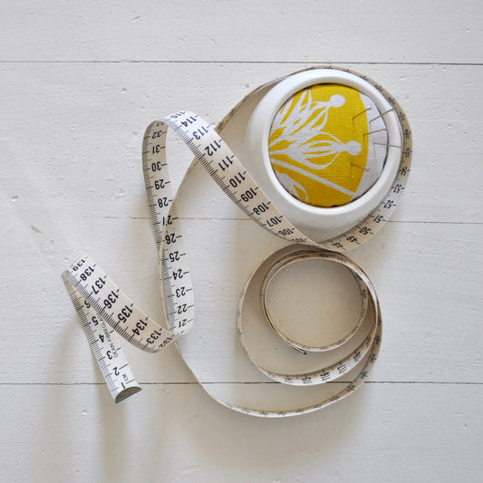 Pinch River ceramic pin cushion featuring Firewheel print in Buddha Gold (colourway 1_v2). Top view. Product image is styled with a measuring tape and pins.